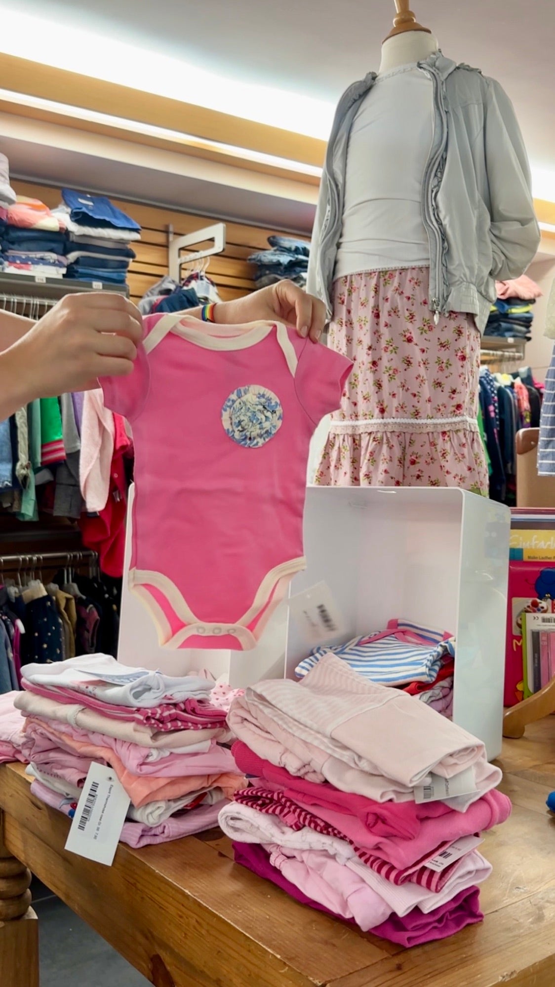 Rosa Babybody wird im Laden hochgehalten, davor gestapelte Babykleidung